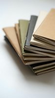 Stack of fanned kraft paper notebooks in earthy tones arranged on white surface, showcasing natural textures and minimalist stationery design.