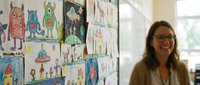 Young female teacher with glasses smiling in classroom with colorful student artwork and drawings displayed on wall behind her.