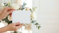 Hands holding blank white card with fresh flowers and greenery in soft focus background, suitable for greeting cards, invitations, and promotional materials.