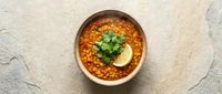 Hearty lentil soup with fresh herbs and lemon slice in ceramic bowl on textured stone surface for healthy cooking and restaurant menu design.