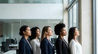 Young diverse women professionals standing in modern office space with large windows, wearing business casual attire, representing teamwork and corporate diversity in workplace environment.