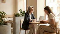 Two professional businesswomen having productive meeting in modern office cafe discussing project collaboration and partnership strategies.