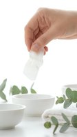 Adult hand with light skin tone holding white facial tissue above ceramic bowl, with fresh eucalyptus leaves and rolled towel nearby for skincare, wellness and spa applications.