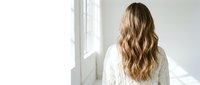 Young blonde woman in white sweater looking out bright window in minimalist bedroom interior with natural morning light and peaceful atmosphere.