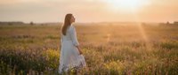 Woman in white dress standing in blooming wildflower field at golden hour sunset, peaceful countryside landscape for lifestyle and nature photography.