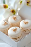 Elegant white macarons decorated with rose gold flakes served in minimalist box, with soft-focused white flowers in background. Luxury dessert styling.