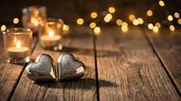 Two silver heart-shaped ornaments on rustic wooden table with warm glowing candles and bokeh lights creating romantic Valentine's Day atmosphere.
