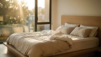 Unmade bed with beige linens and wooden headboard in modern bedroom with large windows and warm sunlight streaming through, suitable for home decor and lifestyle content.