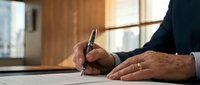 Mature male professional signing document at wooden desk with window background, business contract execution.