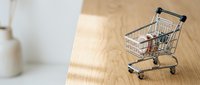 Young woman with long brown hair, wearing casual clothing, holding a miniature shopping cart filled with groceries on a wooden surface, representing retail, consumerism, and online shopping concepts.