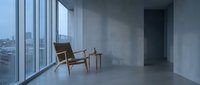 Minimalist interior space with wooden lounge chair and side table near floor-to-ceiling windows overlooking cityscape. Modern loft design for architectural and lifestyle projects.