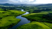 Meandering river flows through vibrant green rolling hills and valleys under dramatic cloudy sky, creating natural curves in pastoral landscape. Aerial perspective.