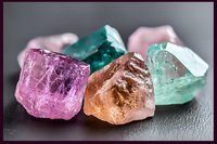 Collection of colorful rough gemstones including pink, green, orange and clear crystals on dark surface for jewelry making and mineral collecting.