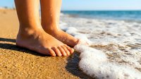 Bare feet of a child standing on sandy beach as gentle ocean wave washes ashore, capturing summer vacation and coastal leisure activities.