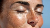 Close-up of young woman with freckles and closed eyes enjoying sunlight on face, showcasing natural beauty and peaceful expression.