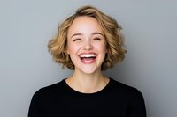 Young Caucasian woman with blonde curly bob hairstyle wearing black top laughing joyfully against gray background, showing genuine happiness and positive emotions.