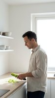 Young Caucasian man in linen shirt chopping leeks on wooden cutting board in bright minimalist kitchen with open shelves.