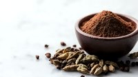 Ground cardamom powder in brown ceramic bowl with scattered whole cardamom pods and spices on white marble surface, macro food photography.