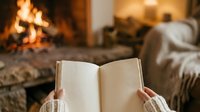 Unrecognizable person holding open book with fireplace and cozy interior in soft focus background, reading by warm fire.