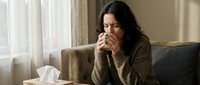 Woman with long dark hair and olive complexion, appearing to be in her 30s, sitting on a beige couch wearing a brown knit sweater, holding tissues to her face while coughing or sneezing indoors.