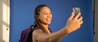 Young African American woman with braided hair taking selfie with smartphone, smiling happily in bright natural light against blue wall background.
