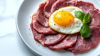 Sliced smoked beef carpaccio arranged on white plate topped with sunny side up egg, fresh basil leaves and cracked black pepper, close up view.