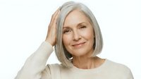 Mature woman with gray hair wearing beige sweater, touching head with hand, smiling at camera against white background. Portrait for healthcare, wellness, aging, lifestyle.