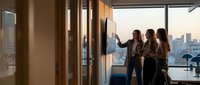 Professional businesswomen collaborating at interactive whiteboard in modern office with city skyline view during golden hour meeting.