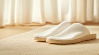 White comfortable home slippers on beige carpet near curtains in morning sunlight. Cozy minimalist lifestyle concept for relaxation and comfort.