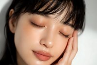 Close-up portrait of Asian woman with closed eyes and natural makeup, resting her face on hand, showcasing warm-toned eyeshadow and matte lip color.