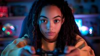 Young African American woman with dreadlocks illuminated by vibrant neon lights holds gaming controller, focused expression while playing video games in dark room with bokeh background.