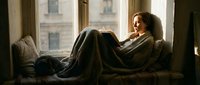 Young woman relaxing on couch wrapped in cozy blanket by window with natural light creating peaceful morning atmosphere for wellness and comfort concepts.