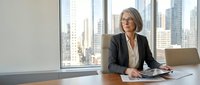 Middle-aged woman with gray hair and glasses wearing dark blazer and white blouse, seated at desk with tablet in modern high-rise office with city skyline view through floor-to-ceiling windows.