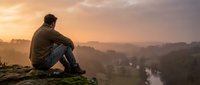 Man sitting on rocky cliff overlooking misty valley at sunrise, contemplating nature landscape with river below, suitable for wellness and lifestyle content.