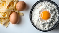 Fresh raw egg yolk in flour bowl with uncooked pasta ribbons and whole eggs on white surface, ingredients for homemade Italian pasta preparation.