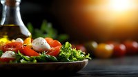 Fresh Mediterranean salad with mozzarella, cherry tomatoes and olive oil in rustic bowl against dark background with golden sunlight, perfect for food blogs.