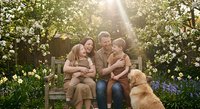 Happy Caucasian family with golden retriever dog sitting on wooden bench in blooming spring garden with sunlight filtering through trees and flowers.