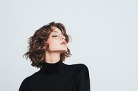 Young Caucasian woman with short wavy brown hair wearing black turtleneck and glasses poses with eyes closed against white background, expressing confidence and style.