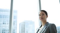 Confident Asian female professional in gray blazer standing by floor-to-ceiling windows overlooking urban skyline, suitable for corporate, business, and leadership concepts.