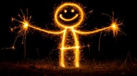 Glowing light painting of stick figure with sparklers creating smiley face and outstretched arms against dark background, captured using long exposure photography.