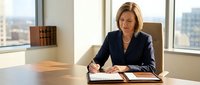 Professional middle-aged Caucasian businesswoman in navy suit writing documents at modern office desk with city view through large windows.