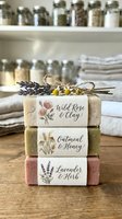 Stacked artisan soaps with natural ingredients including lavender, oatmeal, and clay, decorated with dried flowers and herbs, displayed in a rustic kitchen setting.