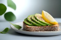 Whole grain toast topped with sliced avocado, lemon wedge, and fresh herbs on white plate for healthy breakfast, brunch, or wellness content.