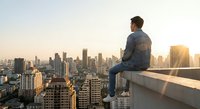 Young businessman sitting on rooftop edge overlooking modern city skyline at golden hour sunset, contemplating future success and urban development opportunities.