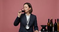 Professional female sommelier in dark suit with medal tasting red wine against pink background, wine bottles and decanter nearby for hospitality industry.