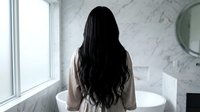 Woman with long dark hair in bathrobe standing by luxury marble bathroom window overlooking modern freestanding tub for spa wellness morning routine.