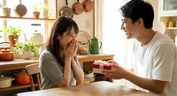 Young Asian couple sharing romantic moment in cozy kitchen as man presents gift box to surprised woman covering face with hands in warm domestic setting.