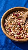 Mixed legumes and beans in wooden bowl with spoon on blue textile background for healthy cooking and nutrition concepts.