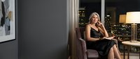 Elegant mature Caucasian businesswoman in black dress sitting in luxury hotel room at night, city lights glowing through window behind her.