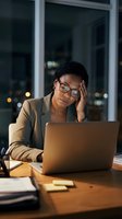 Stressed African American businesswoman working late at office with laptop, experiencing headache and fatigue during overtime hours at corporate workplace.